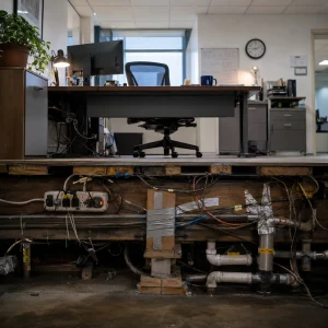 Cutaway view of a tidy office above a floor exposing tangled wiring, duct-taped supports, and patched pipes below.