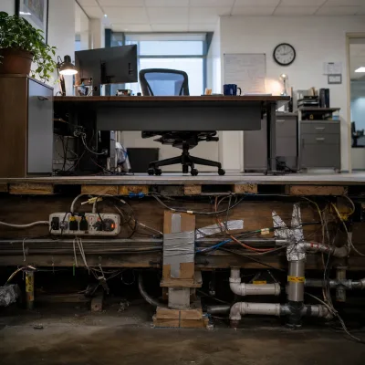 Cutaway view of a tidy office above a floor exposing tangled wiring, duct-taped supports, and patched pipes below.